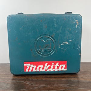 Peut inclure: Une mallette à outils Makita de couleur teal avec un logo rouge et blanc. La mallette a une poignée et deux loquets noirs. Elle présente un emblème circulaire avec les lettres "E.W.".