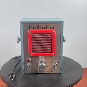 Pode incluir: Um dispositivo eletrônico ByeByeRAT azul e prateado com grade de alto-falante vermelha, botão de controle central numerado, interruptor de energia e seletor Make and Break, fabricado pela Mitsufuji Denko no Japão.