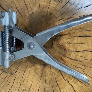 Puede incluir: Una herramienta de metal plateado con un resorte y asas, probablemente para trabajos en cuero o artesanías similares. La herramienta está colocada sobre una superficie de madera con un patrón de vetas natural. El diseño de la herramienta incluye un filo y un mecanismo de resorte.