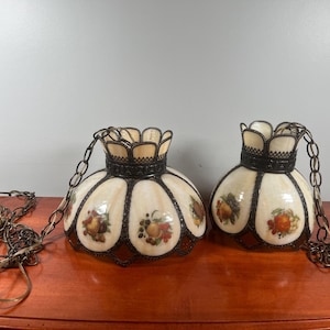 May include: A pair of hanging pendant lamps featuring cream-coloured glass shades with scalloped edges and dark metal frames. Each shade is decorated with colourful fruit illustrations and is suspended by a matching dark metal chain for ceiling mounting.