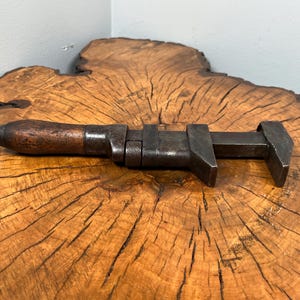 May include: An antique adjustable wrench with a wooden handle and a dark metal body. The wrench is lying on a wooden surface with a natural grain pattern. The tool appears to be well-used and shows signs of age.