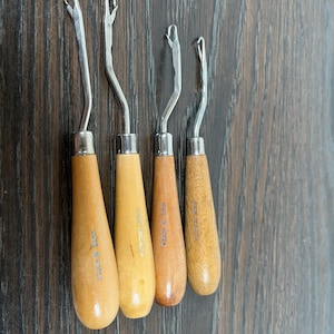Peut inclure: Quatre crochets avec des manches en bois et des crochets en métal. Les manches sont de différentes nuances de brun clair et de beige. Le texte "MADE IN KOREA" est imprimé sur les manches. Les crochets sont disposés sur une surface en bois foncé.