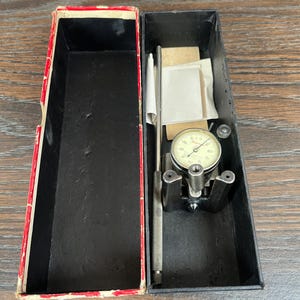 May include: An open black box with a red and white trimmed lid, containing a dial indicator and accessories. The dial indicator has a white face with black markings and a needle. The box is on a wooden surface.