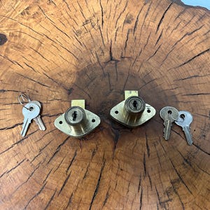 May include: Two vintage brass cabinet locks with keys. Each lock has a diamond-shaped faceplate and a keyhole. The keys are silver-coloured. The locks and keys are on a wooden surface with a natural grain pattern.