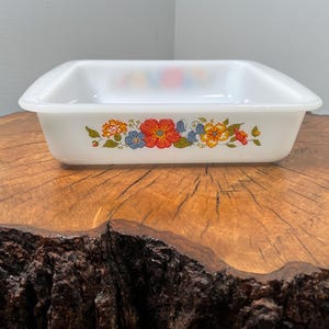 Puede incluir: Fuente rectangular blanca para hornear con diseño floral. La fuente presenta un colorido patrón de flores naranjas, azules y amarillas con hojas verdes. La fuente está sobre una superficie de madera.