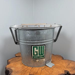 Puede incluir: Un cubo de metal galvanizado con un escurridor de madera. El cubo tiene una etiqueta rectangular con el texto "GW METAL". El cubo está sobre un tocón de árbol de madera.