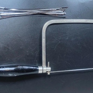 Puede incluir: Una sierra de calar de metal con un mango negro y una hoja, junto con una pequeña pila de hojas de sierra plateadas. Las herramientas están sobre un fondo oscuro, probablemente para la fabricación de joyas o manualidades.