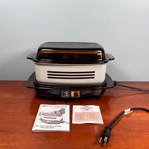 May include: A vintage West Bend slow cooker. It has a black base and lid, and a cream-coloured body with black stripes. Includes a user manual and a power cord. The appliance is on a wooden surface.