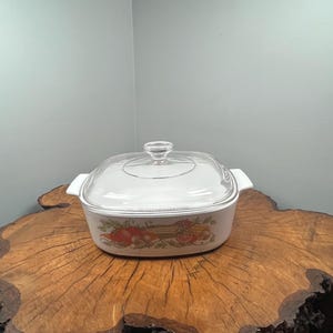 Op de afbeelding: Witte vierkante ovenschaal met een doorzichtig glazen deksel. De schaal heeft een kleurrijk groenteontwerp aan de zijkant. De schaal staat op een boomstronk.