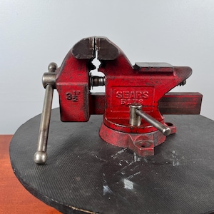 May include: A red Sears 5173 bench vise with a 3.5-inch jaw capacity. The vise is made of metal and has a handle for tightening. It is sitting on a black circular surface.