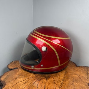 May include: A red motorcycle helmet with gold stripes and a clear visor. The helmet is sitting on a wooden surface.
