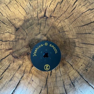 May include: A black Yashica-8 spool with gold lettering and a number 2 in a circle. The spool is centered on a textured, brown wooden surface with visible cracks and growth rings.