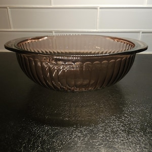 May include: A clear glass bowl with a ribbed design and a brown tint. The bowl is empty and sitting on a black surface.