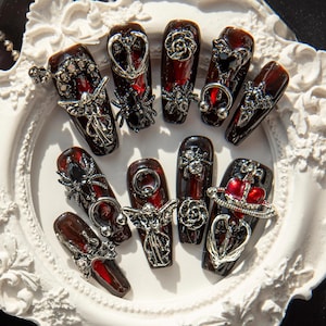 May include: A set of ten red and black gothic-style press-on nails with silver accents. Each nail features a different design, including crosses, angels, and spiders.