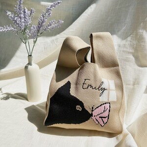 May include: A beige tote bag with a black cat design and a pink butterfly. The bag has the name "Emily" embroidered on it.