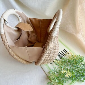 May include: A woven straw handbag with a beige interior and white handles. The bag has a circular handle design and a tan leather tag. The bag is open, revealing the interior. The bag is sitting on a white surface.