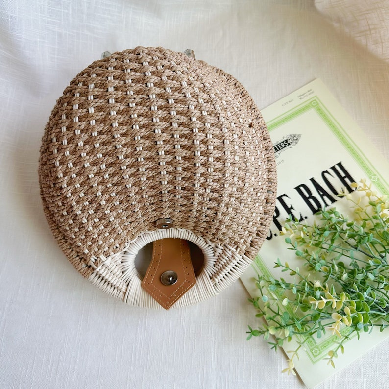 May include: A round, woven handbag in beige and white, featuring a woven pattern. The bag has a leather strap and a button closure. The bag is approximately 10 inches in diameter.