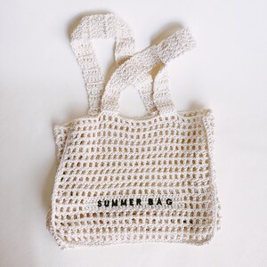 May include: Cream-colored, open-weave crochet tote bag with two shoulder straps. The bag features the text "SUMMER BAG" in black block letters. The bag is made of a thick, textured yarn.