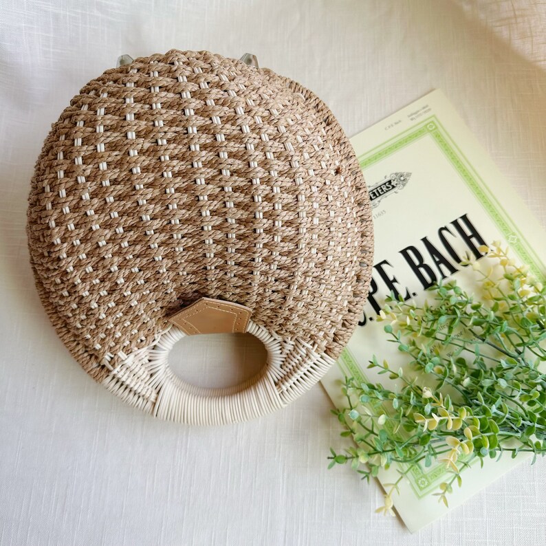 May include: A round, woven handbag in natural tones of beige and white, featuring a circular handle and a small leather accent. The bag is placed on a white surface next to sheet music and greenery.