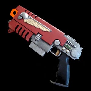 May include: A red and silver prop blaster featuring a prominent silver skull emblem with cream-colored wings on the side. The device includes a black pistol grip, a small digital display screen, circular buttons, and an orange loop at the top rear.