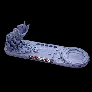 May include: A light purple 3D printed display stand featuring a textured organic sculpture on one end, a circular platform on the other, and five small circular indentations. A plaque with the text RESIDENT EVIL and a red umbrella logo is attached.
