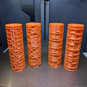 Puede incluir: Cuatro objetos cilíndricos naranjas con superficies texturizadas, posiblemente impresos en 3D. Los cilindros tienen diferentes patrones, incluyendo texturas de rejilla y orgánicas. Están dispuestos sobre una superficie oscura, probablemente una base de impresora.