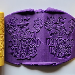 Può includere: Mattarelli gialli con un disegno di mago e le parole "Dungeon Master" sono esposti accanto a un'impronta di argilla viola dello stesso disegno. Il mago tiene un dado.