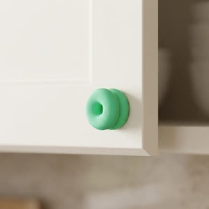 Può includere: Un pomello per armadietto color verde menta dalla forma circolare, simile a una ciambella, con dettagli ad anelli concentrici sulla superficie. Il pomello è montato su un'anta bianca, fungendo da maniglia per aprire e chiudere il mobile.