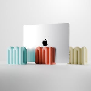 Könnte beinhalten: Ein silberner Laptop mit schwarzem Apple-Logo wird von drei Sätzen farbiger, abstrakter, geschwungener Formen flankiert. Die Formen sind in Hellblau, Koralle und Creme gehalten. Das Design ist modern und minimalistisch.