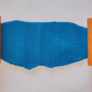 Könnte beinhalten: Ein strukturiertes, leuchtend blaues rechteckiges Objekt mit gewelltem oberem Rand, flankiert von zwei orangefarbenen zylindrischen Objekten. Das blaue Objekt hat eine raue, zerknitterte Oberfläche. Die orangefarbenen Zylinder haben eine rindenartige Textur. Die Objekte sind vor weißem Hintergrund.