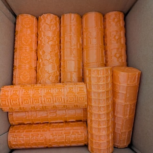 Könnte beinhalten: Eine Sammlung orangefarbener zylindrischer Objekte mit verschiedenen strukturierten Mustern, darunter geometrische und florale Designs. Die Objekte sind in einem Karton angeordnet und zeigen ihre einzigartigen Oberflächendetails und Formen.