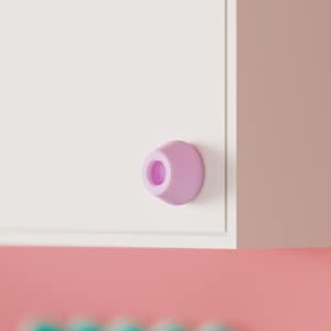 Pode incluir: Um puxador de armário de formato cónico na cor lavanda claro com uma abertura circular no centro, montado numa superfície plana de porta de armário retangular branca. O puxador apresenta um acabamento mate liso e um design cónico.