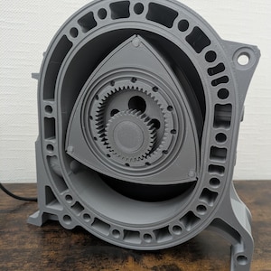 Puede incluir: Modelo gris impreso en 3D de un motor rotativo, que muestra engranajes internos y carcasa del rotor. El modelo es detallado con componentes visibles y un diseño circular, ideal para entusiastas del automóvil o fines educativos.