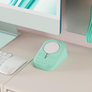 Può includere: Un caricatore wireless verde menta, un supporto per telefono abbinato e un supporto con bordi ondulati sono esposti su una scrivania bianca. Sono visibili anche una tastiera, un mouse e un monitor per computer bianchi.