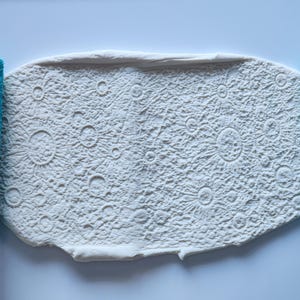 May include: A white, textured surface with a moon crater design is flanked by two turquoise rolling pins. The rolling pins have a matching moon crater pattern. The design is ideal for baking or crafting.