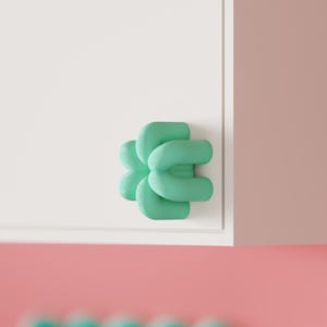 Puede incluir: Un pomo de armario de color verde menta con una forma redondeada de cuatro lóbulos similar a un trébol. El pomo está montado sobre una superficie blanca plana. El diseño consta de cuatro segmentos tubulares lisos e interconectados.