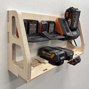 May include: A wooden power tool battery organizer with multiple slots for holding batteries. The organizer is made of light-colored plywood and features a shelf design. Several black and orange batteries are inserted into the slots.