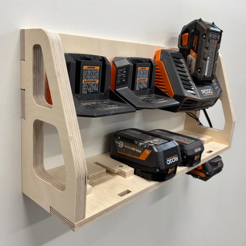 May include: A wooden power tool battery organizer with multiple slots for holding batteries. The organizer is made of light-colored plywood and features a shelf design. Several black and orange batteries are inserted into the slots.