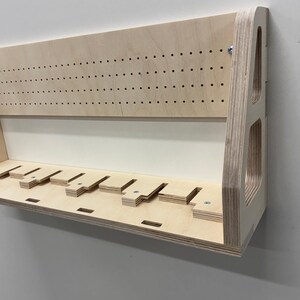 May include: A light-colored wooden wall-mounted shelf with multiple slots and a pegboard-style top section. The shelf has a simple, modern design, with visible wood grain and a clean, minimalist aesthetic. It is mounted on a white wall.