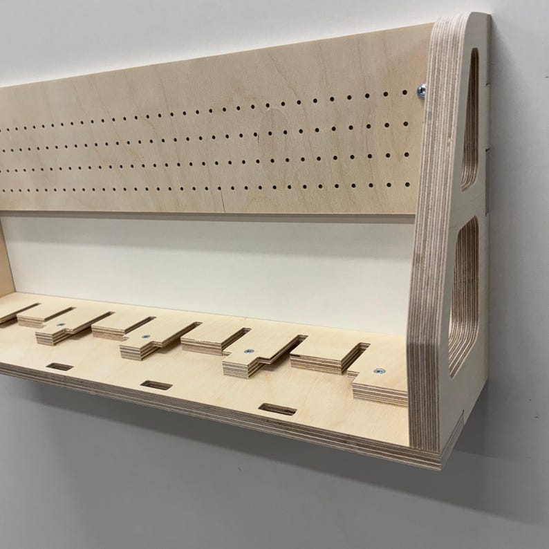 May include: A light-colored wooden wall-mounted shelf with multiple slots and a pegboard-style top section. The shelf has a simple, modern design, with visible wood grain and a clean, minimalist aesthetic. It is mounted on a white wall.