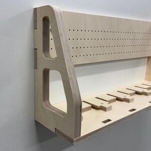 May include: A light-colored wooden wall shelf with a pegboard back and a lower shelf with several slots. The shelf has a modern design with cut-out details and is mounted on a white wall.