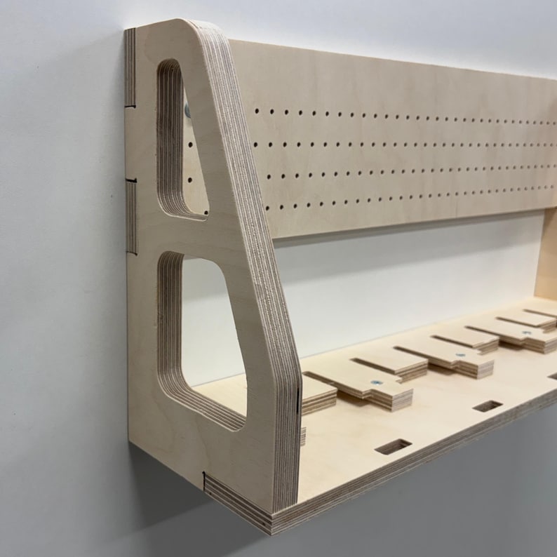May include: A light-colored wooden wall shelf with a pegboard back and a lower shelf with several slots. The shelf has a modern design with cut-out details and is mounted on a white wall.