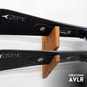 May include: Black skis branded "Atomic" are displayed on a wooden ski rack. The rack is made of light brown wood and is mounted on a white wall. The text "CRÉATIONS AVLR" is visible in the bottom right corner.
