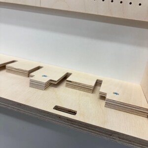 May include: Close-up of a wooden shelf with several cut-out sections and small wooden blocks. The shelf is made of light-colored plywood and has a clean, minimalist design. Small screws are visible, securing the wooden blocks.