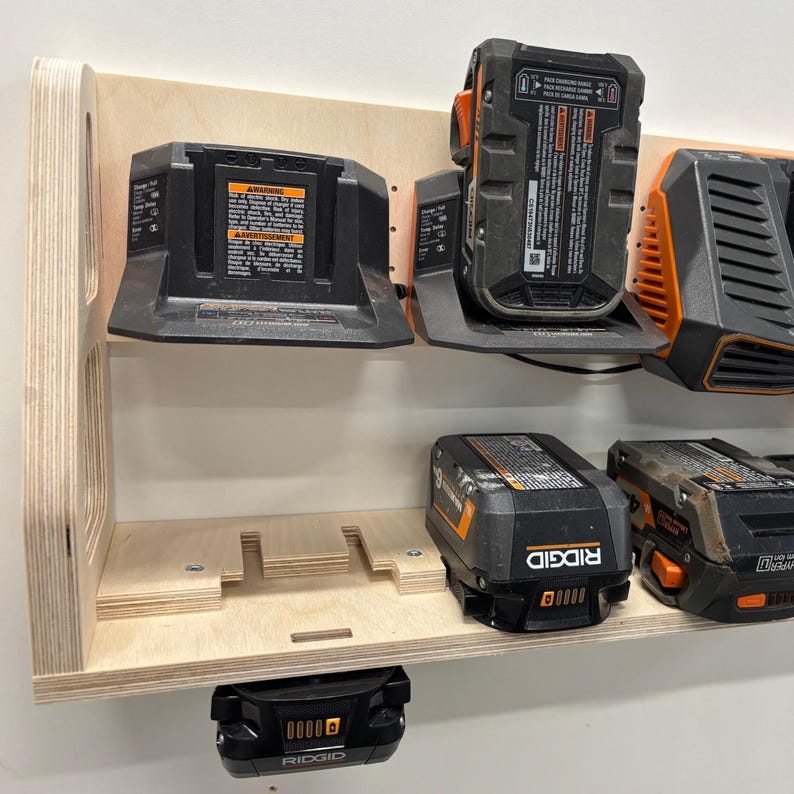 May include: A wooden wall-mounted power tool battery organizer. The organizer holds multiple black and orange power tool batteries and chargers. The wooden shelf is made of light-colored plywood and is mounted on a white wall.