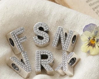 May include: Six silver-toned letter charms featuring the letters F, S, N, V, R, and X, each encrusted with small clear stones. The charms are arranged on a piece of off-white fabric next to a single yellow and purple pansy flower blossom.
