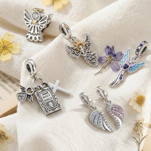 May include: A collection of silver charms including an angel with a gold halo, a sun-adorned angel, a winged figure with blue and pink enamel, a cross and Holy Bible charm set, and a pair of angel wing charms accented with blue and pink crystals.