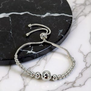 May include: A silver bracelet with a sliding clasp. The bracelet is made up of small, round beads and a larger central bead. The adjustable closure allows for a custom fit. The jewellery is displayed on a black and white marble surface.