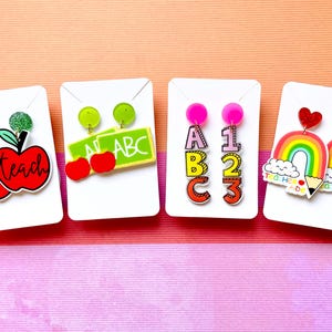May include: Four pairs of colourful teacher-themed earrings. The first pair features red apples with the words "tea teach". The second pair features green apples and a green chalkboard with the letters "ABC". The third pair features colourful letters spelling "ABC" and numbers "123". The fourth pair features rainbow-coloured rainbows with clouds and pencils that say "Tea Her Aide".
