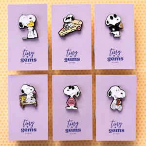 May include: Six enamel pins featuring the character Snoopy in various poses, each mounted on a light purple card labelled tiny + gems store. The pins depict Snoopy eating, driving a pizza slice, wearing sunglasses, sitting on a bench, wearing a pink jumper, and holding a mug.